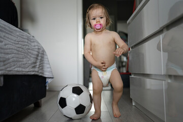 One female toddler caucasian in diapers running in the house with soccer ball Early child development learning and playing concept - obrazy, fototapety, plakaty