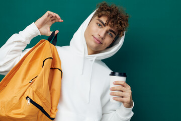 a nice man stands on a green background in a white hoodie with an orange backpack on his back and holding a glass in his hand looks at the camera