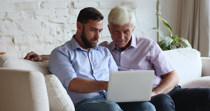 Happy grown son teaching older father to use online app on laptop, sharing computer, speaking, telling, explaining Internet service, application work, pointing at display, laughing
