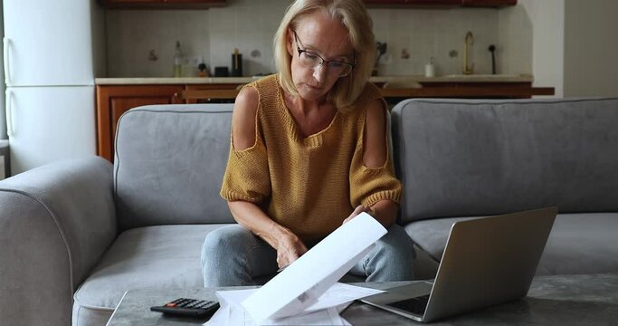 Serious mature homeowner woman in glasses calculating monthly expenses, budget, reviewing paper bills, checking invoices, using online payment app on laptop, typing, paying taxes, insurance fees