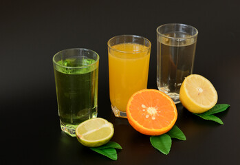 Three glasses with different citrus juices and next to each of them are halves of lime, lemon and orange on a black background. Close-up.