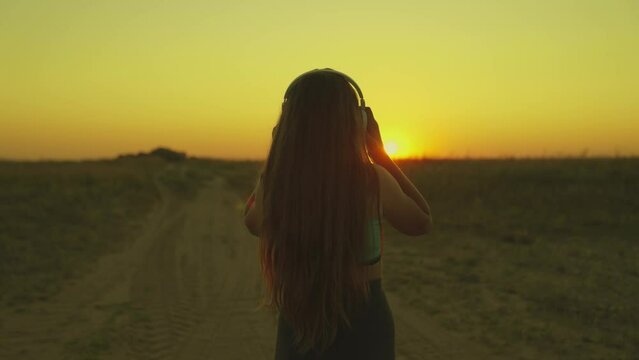 young girl runs across field sunset headphones. silhouette girl rays sunlight listens music. healthy image women health. morning run outdoors cardio. exercises weight loss. long-haired sun girl.