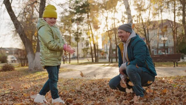 father with little child kid throws autumn leaves sunlight glare. autumn sunset park. laughing happy family. child has fun playing with dad leaves .kid plays game with parent nature. flying leaves.