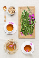 Obraz na płótnie Canvas Herbal tea from kipreya leaves in white cups on linen fabric table background, fireweed green leaves and flowers on wooden tray. Flavored herbal tea from wild plants, healing hot beverage.
