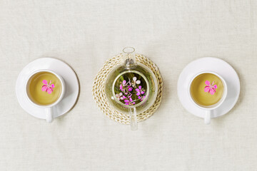 Obraz na płótnie Canvas Herbal tea from kipreya leaves in white cups on linen fabric table background, fireweed green leaves and flowers on wooden tray. Flavored herbal tea from natural plants, healing hot beverage.