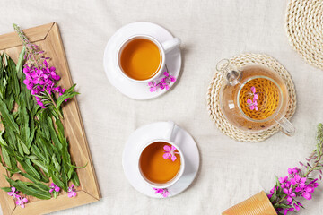 Obraz na płótnie Canvas Herbal tea from kipreya leaves in white cups on linen fabric table background, fireweed green leaves and flowers on wooden tray. Flavored herbal tea from natural plants, healing hot beverage.