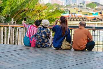 Back view portrait of attractive group of Asian friends or travellers using a smartphone or mobile phone take photo at riverside on sunny day during weekend trip. Attractions and travel concept.