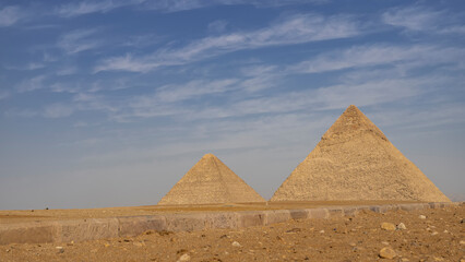 Fototapeta na wymiar Two great pyramids of Cheops and Chephren on a background of blue sky and clouds. In the foreground is the sandy-rocky soil of the Giza plateau, a road curb. Egypt