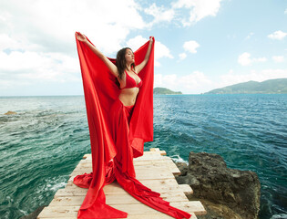 Young woman with red skirt and fabric like a wings. female model near sea. Summer time.