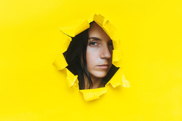 Close-up portrait of a caucasian young woman looking through a hole in yellow paper. An incredulous...