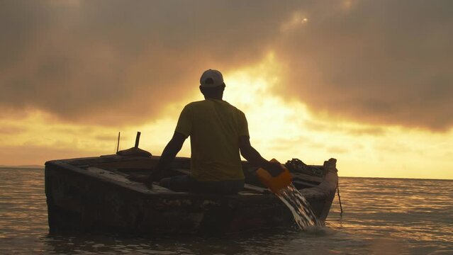 Silhouette Of Fisherman Sitting In Small Fishing Boat Throwing Water Out Of Small Boat During Gorgeous Golden Hour Ocean Sunset In Vilanculos Bazaruto Archipelago, Mozambique, Africa.