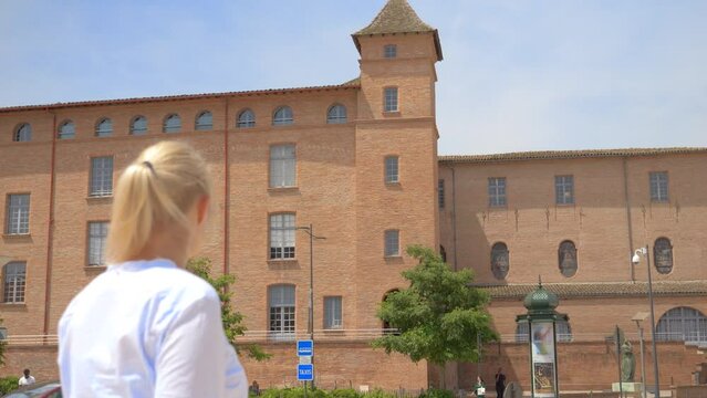 A blonde Caucasian woman walks down the street in front of a big old red stone building with lots of windows