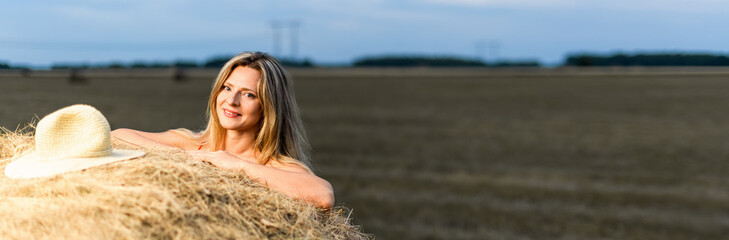 Girl and haystack. Smiling blonde woman leaning on a haystack in a field at sunset. Harvest...