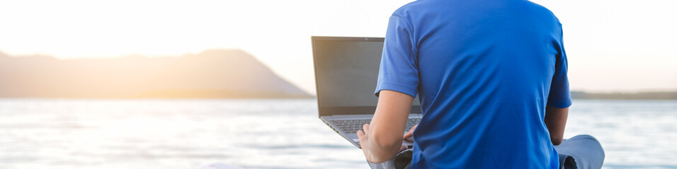 young hipster digital nomad man sitting on wooden pier at sea working on internet remotely at sunset - Traveling with a computer - Online dream job concept - Selective Focus.