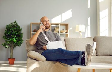 Relaxed man with broken arm having fun talking on phone with friends during rehabilitation at home. Cheerful bald young man in immobilizer on injured arm sitting on sofa throwing legs on coffee table. - obrazy, fototapety, plakaty