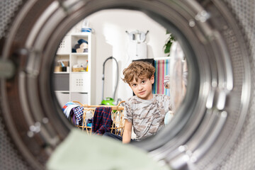 Fototapeta na wymiar Small cheerful boy interested in the operation of the device puts clothes into the drum of the washing machine, helps his mother in household duties, resourceful, hard-working child.