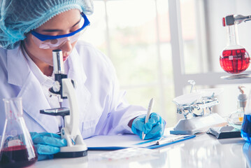 Fototapeta premium Asian Woman scientist hand write note research in medical lab look at science microscope medical test. Researcher biology chemistry. Asian Female technician laboratory using microscope blood test