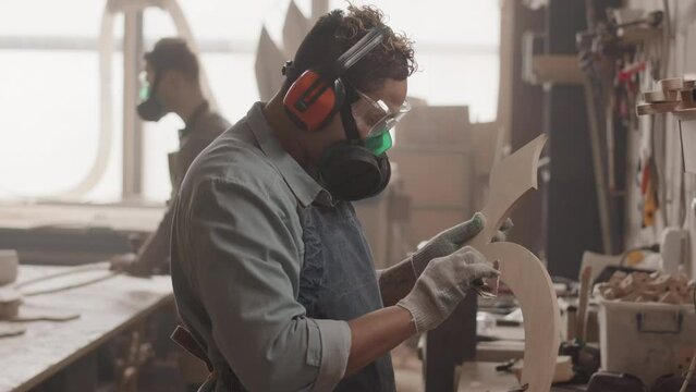 Slowmo of female carpenter in earmuffs and dust mask polishing carved piece of wood while making furniture in workshop with colleague