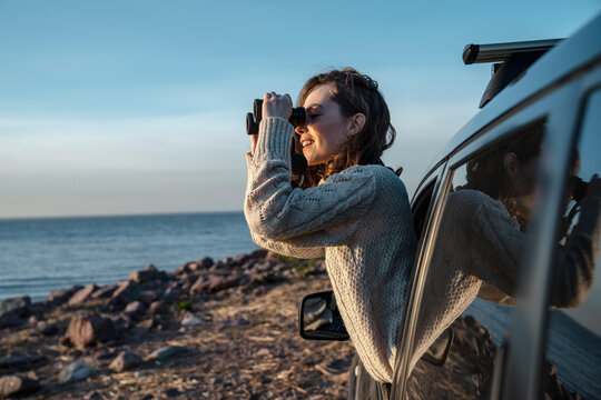 Smiling young woman looking through binoculars leaning out of car