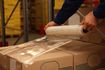 Worker wrapping boxes in stretch film at warehouse, closeup - obrazy, fototapety, plakaty