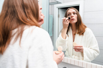 Obraz na płótnie Canvas A young woman in a white bathrobe puts makeup on her face with a brush. Spacious bright bathroom with mirror