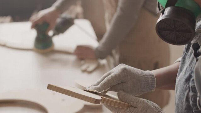 Slowmo of two woodworkers in dust masks polishing carved wooden items using angle grinder, making furniture in carpentry workshop