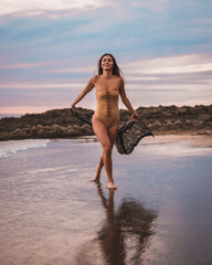 A brunette woman having fun with a swimsuit and a pareo walking along the beach in summer at sunset
