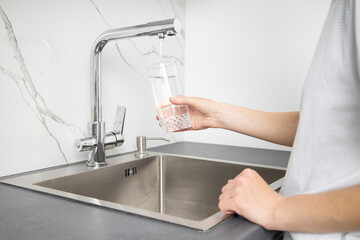 Young woman holding a glass pours water from a faucet in the kitchen