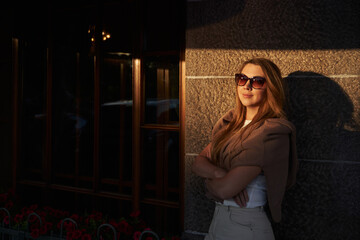 beautiful woman in sunbeams and sunglasses near the building with high windows. city walks in summer, tourism. vacation in the city