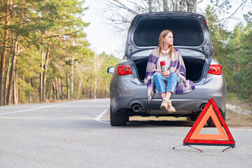 Red emergency triangle with car and woman sitting in the trunk with thermos in the background waiting help. Broken car on the road