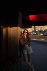 a woman in the sun at the building with red visors on the windows stands leaning against the wall. city walks in summer, tourism. vacation in the city