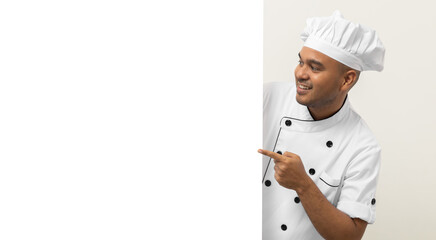 Happy Young handsome asian man chef in uniform looks out from behind an empty whiteboard. Cooking indian man hiding behind big blank billboard for advertising text menu in kitchen and restaurant.