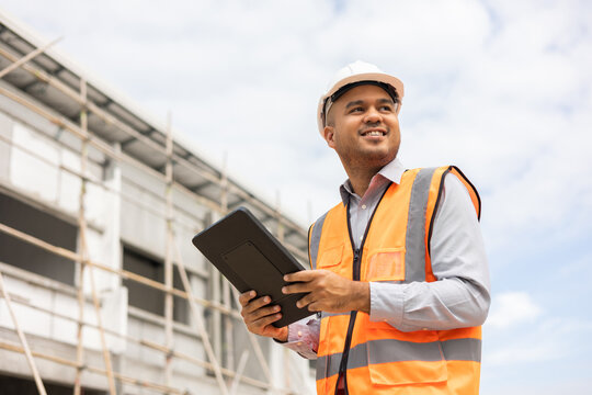 Confident asian engineer man Using tablet for checking and maintenance to inspection at modern home building construction. Architect working with white safety helmet in construction site
