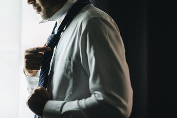 Confident businessman tying or adjust the necktie near window in hotel room in the morning....