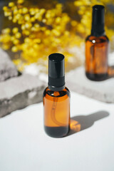 Amber pump bottles with serum, tonic or essential oil on grey concrete podium with yellow flowers. White background with daylight. Beauty concept for face and body care