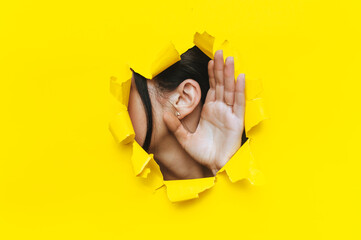 Close-up of a left woman's ear and hand through a torn hole in the paper. Bright yellow background, copy space. The concept of eavesdropping, espionage, gossip and tabloids.