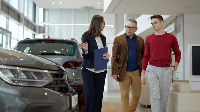 Female manager is talking to male customers and showing new cars in company spbd. 4k Young beautiful woman talks to two men and looks with smiles, shows transport models and walks in light interior