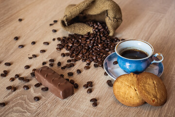 Roasted coffee beans in little bag and cup of coffee with drink on dark wooden surface, nearby lies chocolate and appetizing cookies.