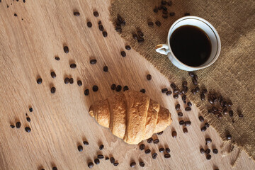 A cup of fragrant black coffee, a croissant and coffee beans on a wooden brown textured table...