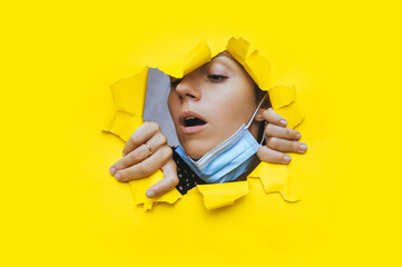 Surprised young woman with open mouth and protective medical mask looks through a torn hole in yellow paper. Concept of protection measures against pandemic, quarantine, coronavirus and covid-19.