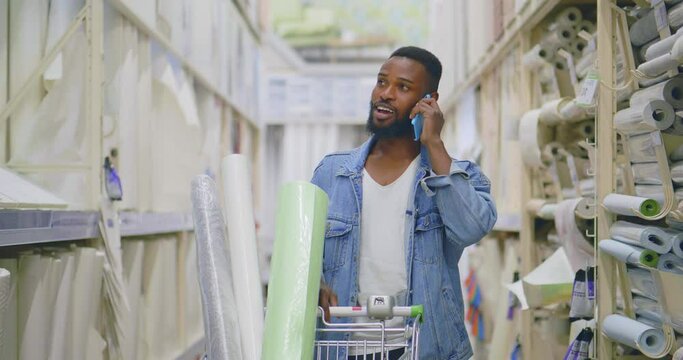 African-American man with shopping cart talk on smartphone walking in diy store. Realtime