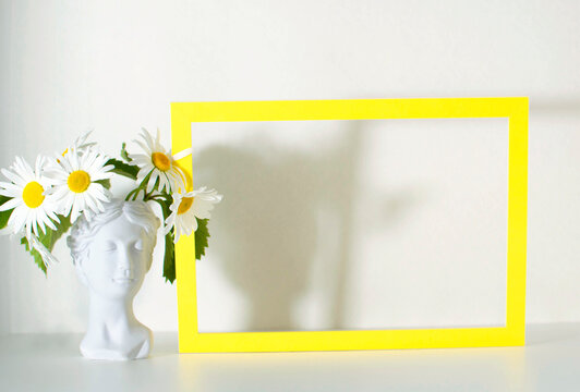A mock-up in a yellow frame with a daisy flower in a vase on a white wall background