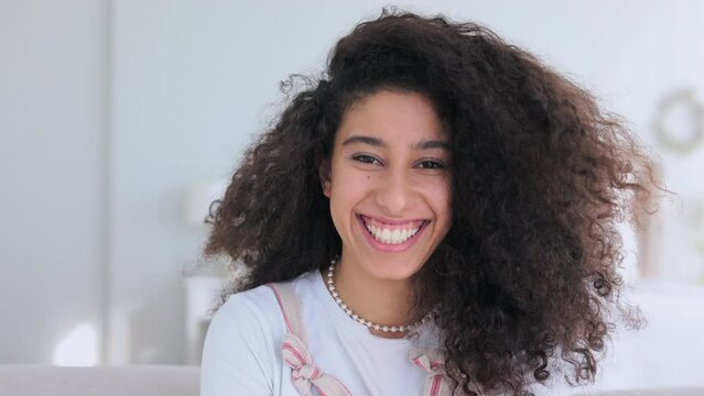 Portrait of young beautiful woman is posing and smiling while sitting on sofa in apartment spbi. 4k Close view of curly Caucasian female looks with toothy smile and laughs, poses for camera in good