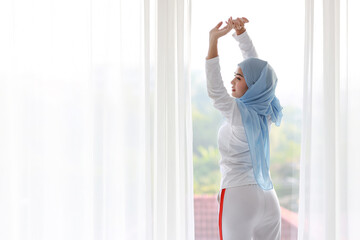 Fototapeta na wymiar Beautiful asian woman wearing white muslim sleepwear, stretching her arms after getting up in the morning at sunrise. Cute young girl with blue hijab standing and relaxing with happy and smiling face.