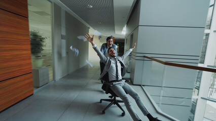 Two crazy businessmen riding office chair and throwing papers up while having fun in lobby of...