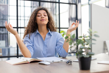 Calm beautiful businesswoman practicing yoga at work, meditating in office with eyes closed.
