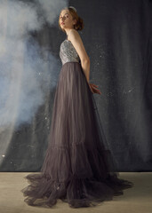 Fototapeta premium a woman in a long evening dress stands against a dark background, posing relaxed in a smoky room, with a crown on her head
