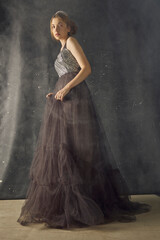Fototapeta premium a woman in a long evening dress stands against a dark background, posing relaxed in a smoky room, with a crown on her head
