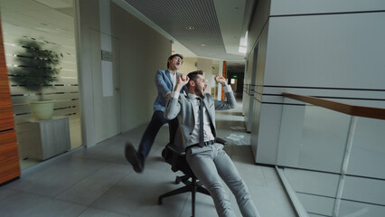 Two crazy businessmen riding office chair and throwing papers up while having fun in lobby of...