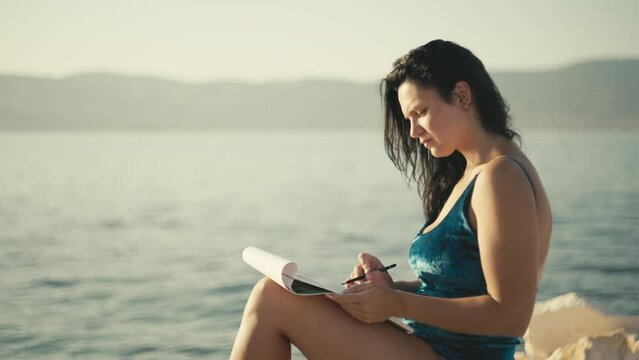 The girl on a stone by the sea draws in the album with a pencil. Beautiful woman is engaged in hobbies in nature.